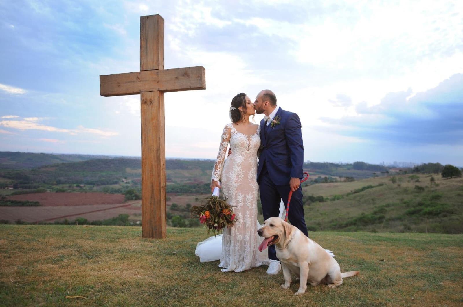 Noivos e cachorro diante da cruz de madeira com vista para o vale